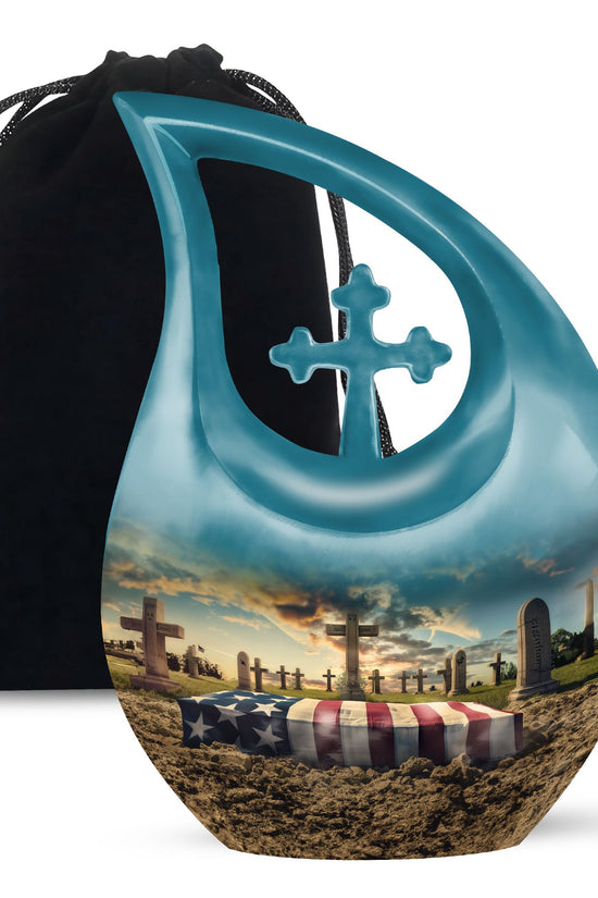 Teal urn with cross design on a black drawstring bag, featuring a scenic design of a cemetery with an American flag.