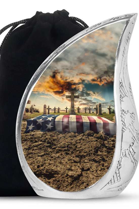 Silver urn with a scenic design of a casket under an American flag and cross, with a black drawstring bag.