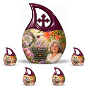 Hummingbird 01 Cross Drop, 10 inch large Urn with 4 Keepsake 3 Inch Urns: glossy mulberry-plum loop above a ruby-throated hummingbird, coral trumpet flowers and rose-lavender garden; fictional Celia Roswick portrait, name and full dates.